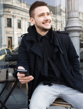 Nice man sitting on chair in steet, texting message on phone, smiling, looking away.